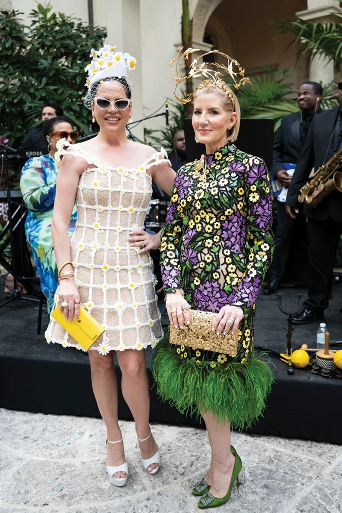 Two women in floral dresses holding clutch purses and wearing elaborate hats.
