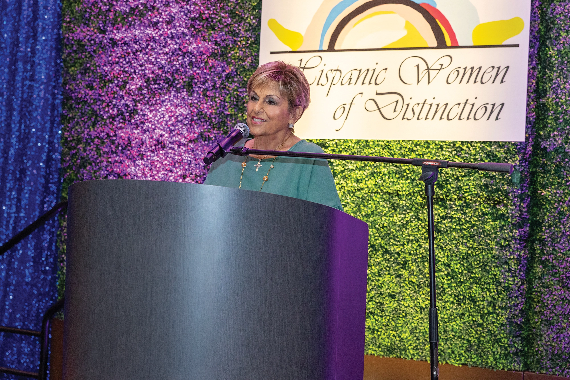 Woman on stage with a microphone, behind a podium, with a plant wall behind her.