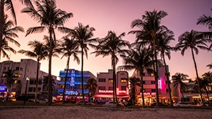 View of palm trees and hotels on sandy beach with pink sunset sky.