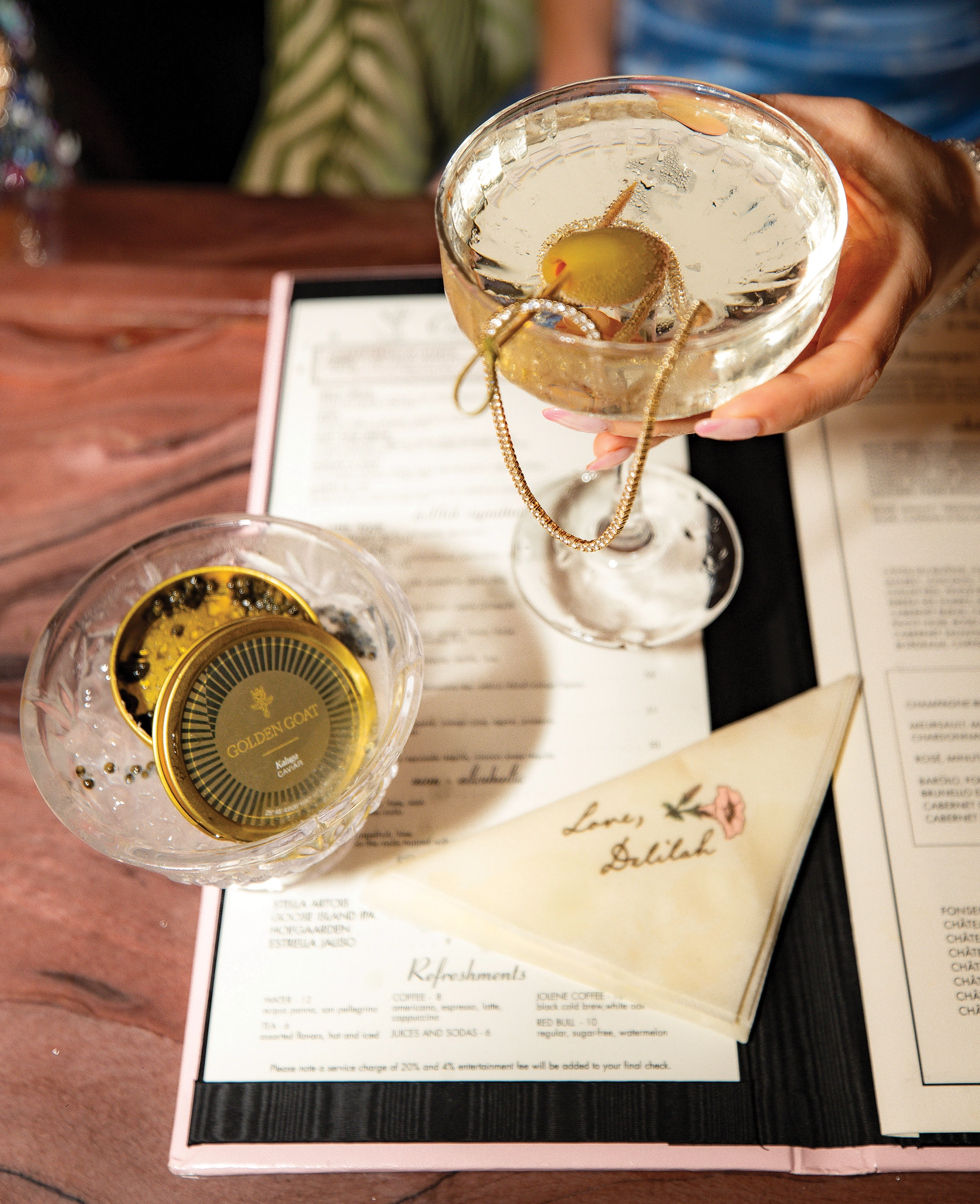 A martini with olive and diamond necklace wrapped around the martini glass, with a tin of caviar, menu and monogrammed napkin underneath.