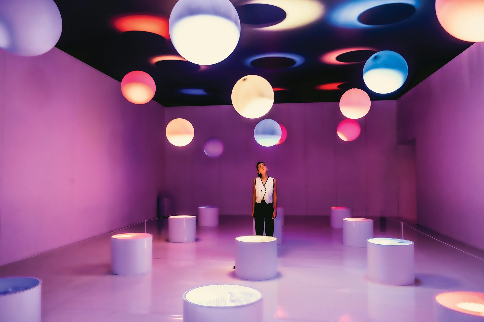 Woman standing in middle of room with colorful balloon art orbs overhead.