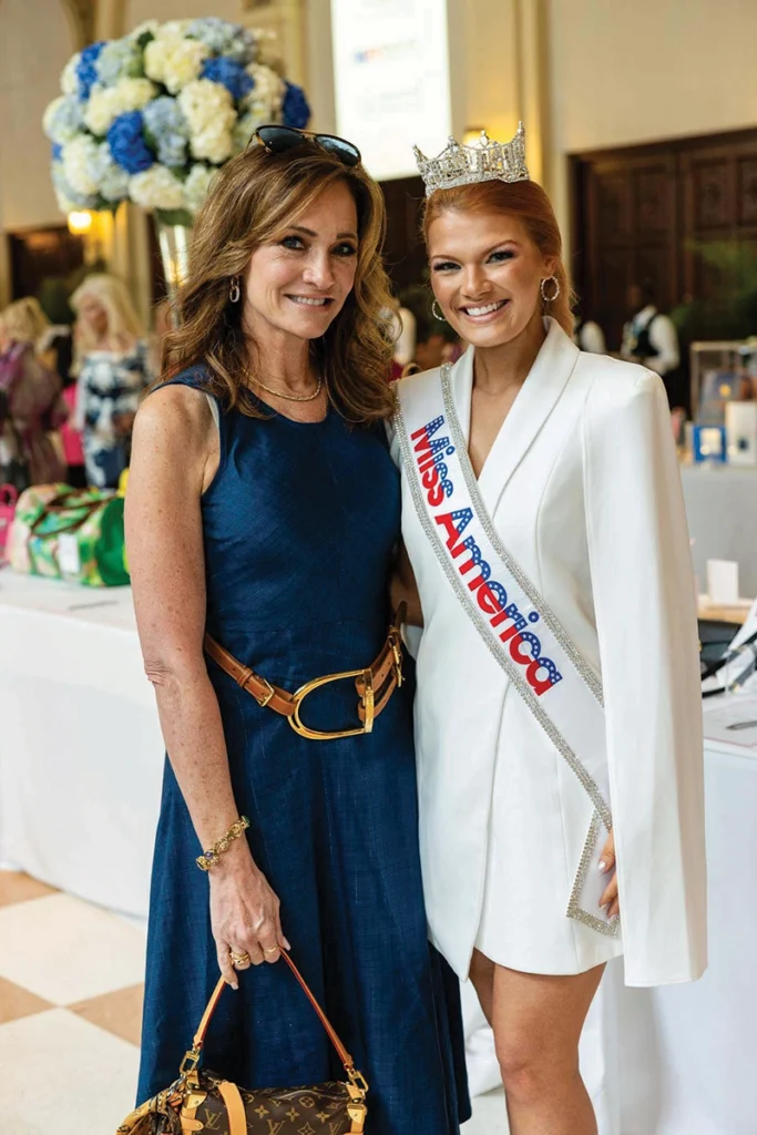 Two women smiling, one on the left wearing beauty pageant sash and tiara.