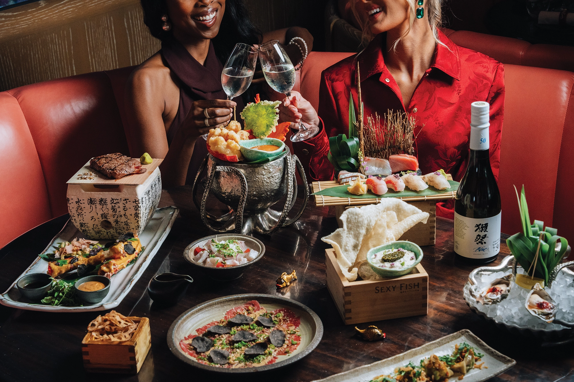 Two women holding glasses with sake at a table with dishes of food and a bottle of sake.