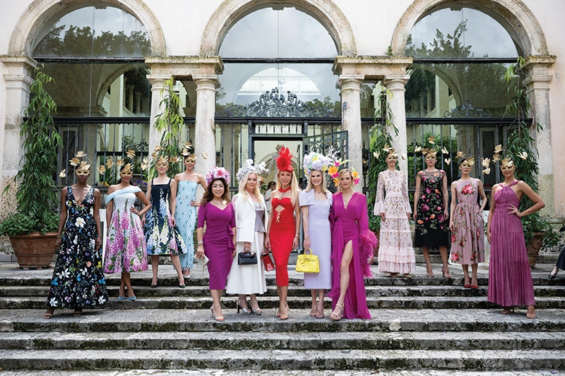 A group of women standing on steps, smiling, wearing elaborate hats and dresses.